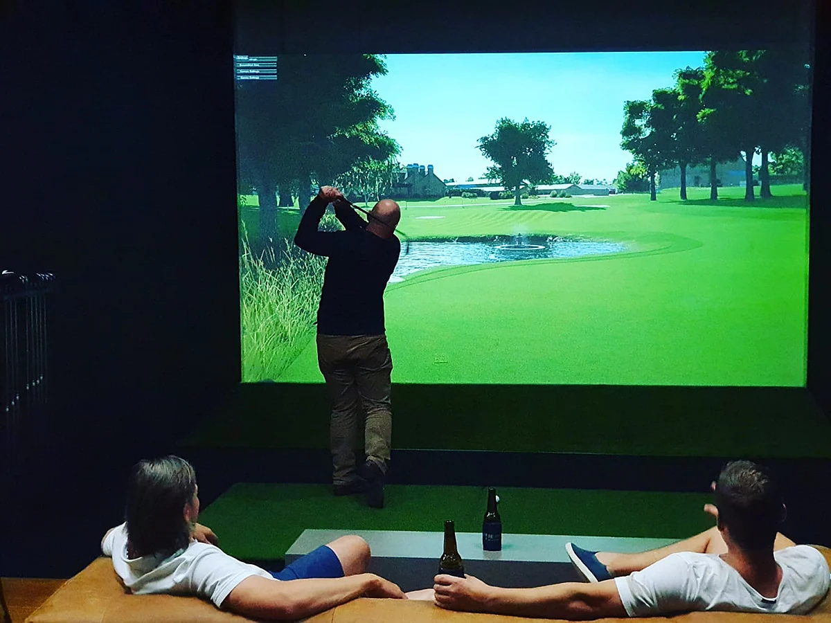 Inside a FOREplay Golf bay: one player mid-swing at the simulator screen while two mates watch from the couch with beers.