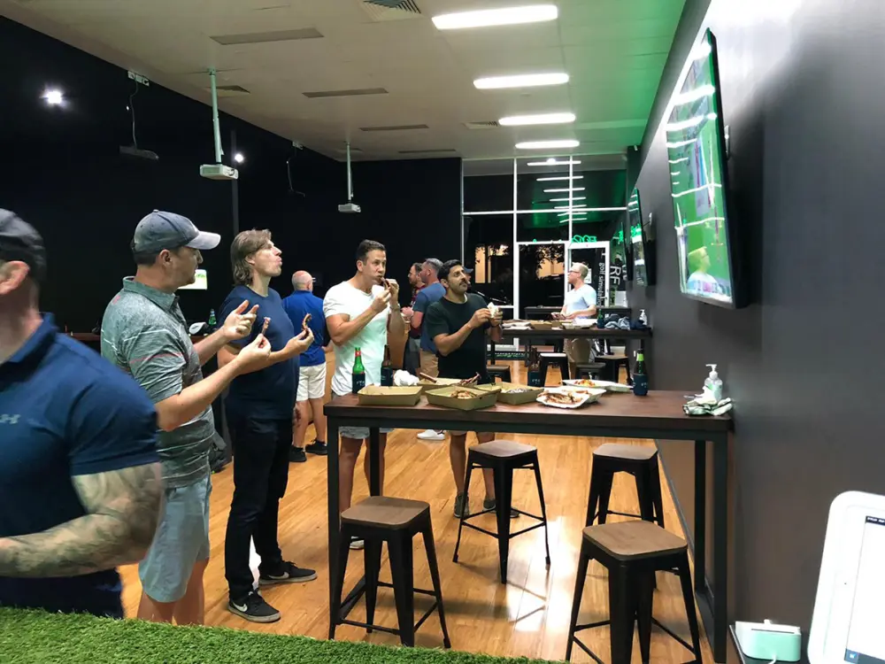 A group standing around a high table with food and beers, golf on the screens above.