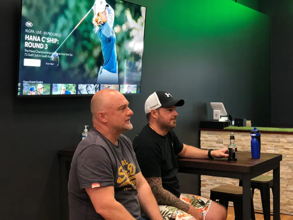 Two players sitting at a high table watching tour golf on the wall-mounted screen.