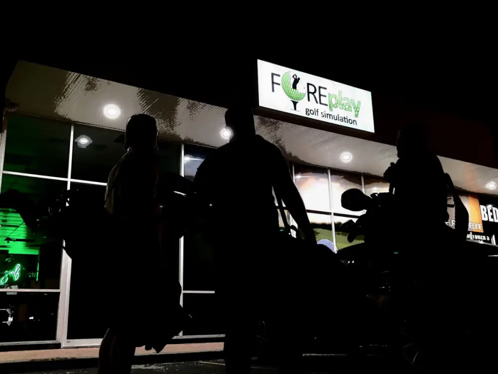 The lit FOREplay Golf shopfront at night with players carrying bags in.